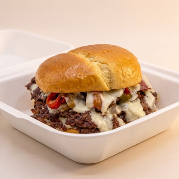 Cheesesteak sandwich with melted cheese, peppers, and onions in a white takeout container.