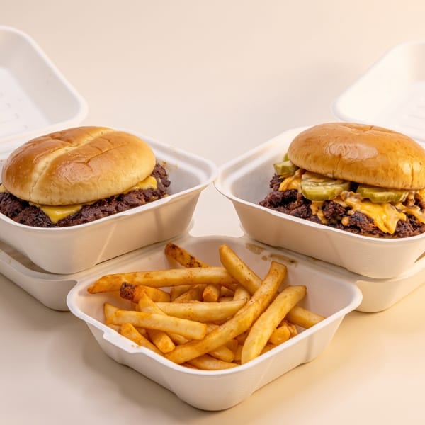 Two cheeseburgers in open takeout containers with a side of french fries in the foreground.