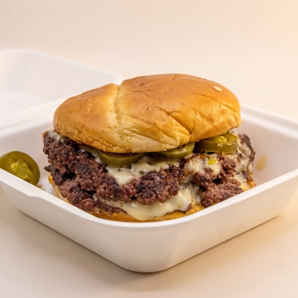 A smash burger with melted cheese and jalapeños in a white takeout container.