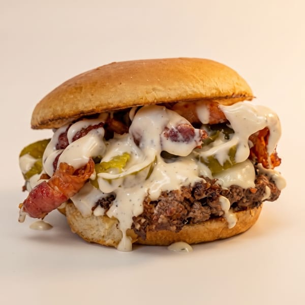 A loaded burger with a beef patty, bacon, pickles, and white sauce on a bun.