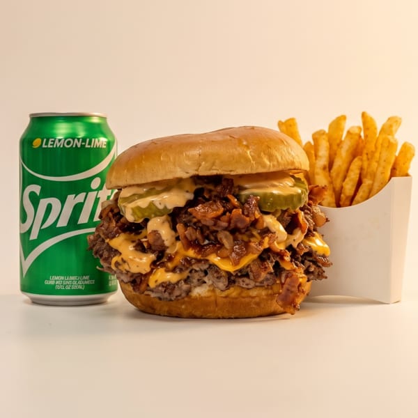A loaded burger with cheese, pickles, and bacon, alongside fries and a Sprite can.